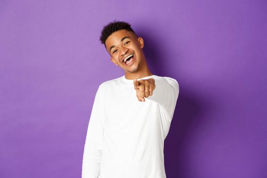 Image of handsome young african-american man in white sweatshirt, pointing finger at camera and smiling, choosing you, standing over purple background.