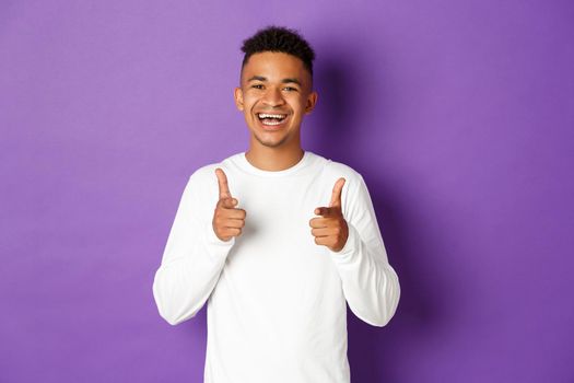Image of cheerful african-american young guy, pointing fingers at camera and smiling, praise good choice, congratulate with something, standing over purple background.
