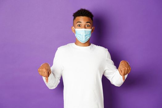 Concept of coronavirus, quarantine and lifestyle. Amazed african-american man in medical mask, pointing fingers down and looking at camera, showing copy space, standing over purple background.