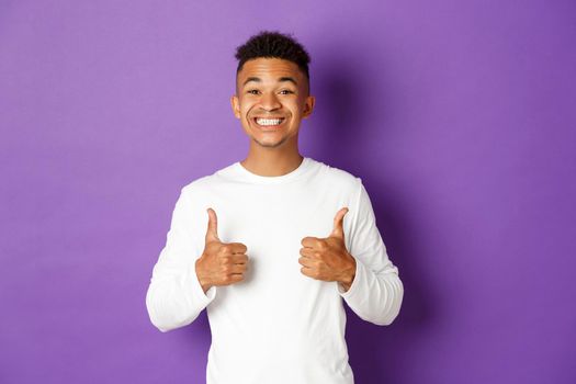 Handsome cheerful man in white sweatshirt, showing thumbs-up in approval and smiling, like and praise something good, standing over purple background.