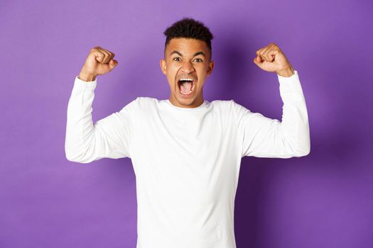 Image of encouraged african-american guy, shouting and looking determined, winning something, achieve goal and celebrating victory, standing over purple background.