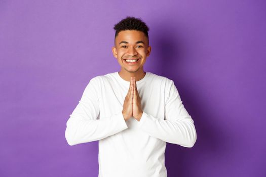 Image of smiling handsome african-american man, thanking for help, holding hands pressed together in pray gesture, looking grateful and happy, standing over purple background.