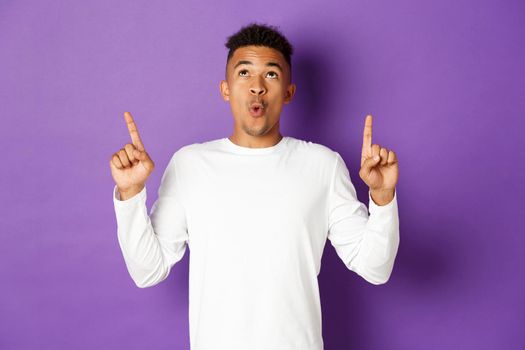 Image of impressed african-american guy in white sweatshirt, pointing and looking up with amazed expression, showing cool advertisement, standing over purple background.