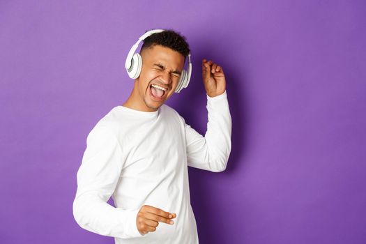 Image of carefree african-american guy dancing, listening music in wireless headphones and singing along, standing over purple background.