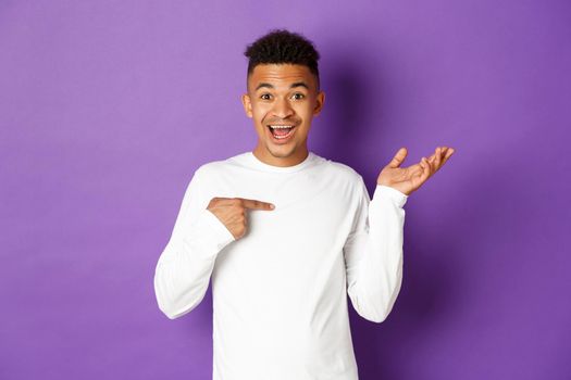 Image of surprised african-american man in white sweatshirt, pointing at himself and raising hand amazed, smiling happy, standing over purple background.