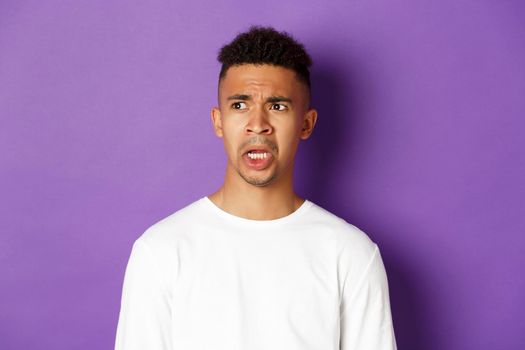 Image of shocked and startled african-american man, drop jaw and looking left at something embarrassing, standing over purple background.