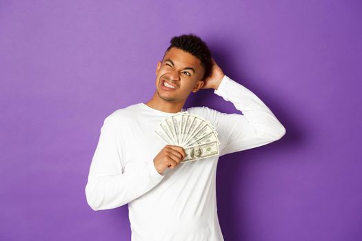 Image of confused african-american man, holding cash and looking at upper left corner puzzled, thinking what to buy, standing over purple background.