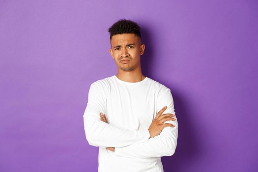 Portrait of skeptical african-american man looking with disbelief, cross arms on chest and frowning doubtful, standing over purple background.