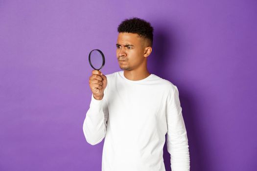 Image of confused african-american man, looking through magnifying glass at something strange, standing over purple background.