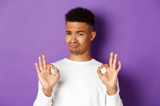 Image of pleased african-american man in white sweatshirt, praise something good, showing okay signs and nod in approval, standing over purple background.