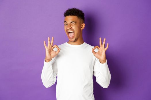 Image of happy and pleased african-american guy, winking and smiling, showing okay signs, recommend good promo offer, like and approve something, standing over purple background.