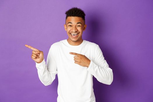 Image of handsome dark-skinned male model in casual outfit, pointing fingers left and laughing at funny advertisement, standing happy over purple background.