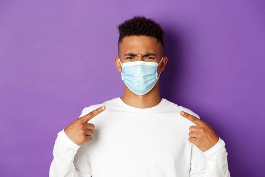 Concept of coronavirus, quarantine and social distancing. Close-up of annoyed african-american man pointing at medical mask, frowning bothered, complaining, standing over purple background.