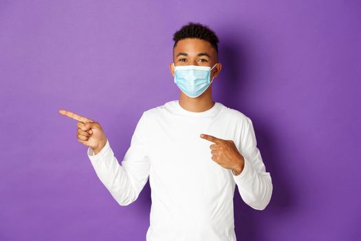 Concept of coronavirus, quarantine and lifestyle. Cheerful african-american man in medical mask showing advertisement, pointing left and smiling, standing over purple background.
