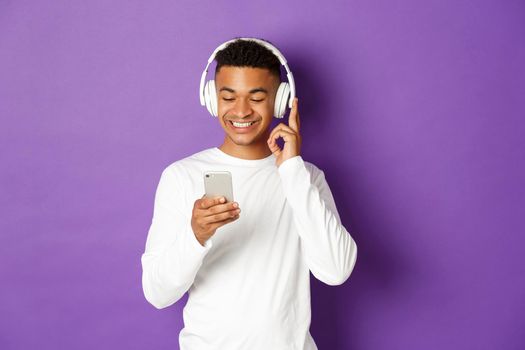 Image of modern african-american guy in white sweatshirt, listening music in headphones and looking at mobile phone, standing over purple background.