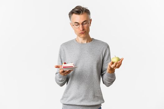 Portrait of thoughtful middle-aged man in glasses, making decision between tasty cake and green apple, standing over white background.