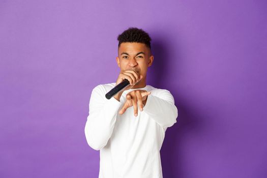 Image of sassy african-american guy singing in microphone, rapping and pointing at camera, standing over purple background.