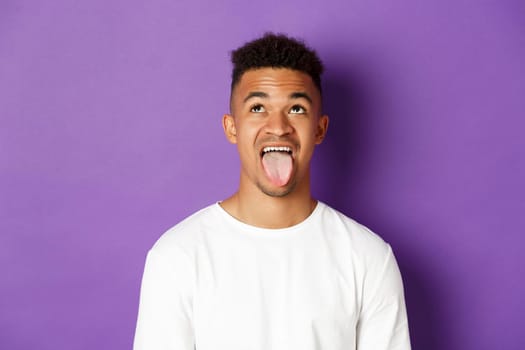 Close-up of silly african-american guy showing tongue, looking up at something lame, standing over purple background.