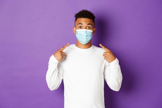 Concept of covid-19, pandemic and social distancing. Image of amused african-american guy, recommend to wear medical mask during coronavirus, standing over purple background.