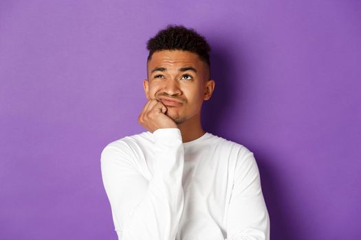 Close-up of indecisive african-american guy, sighing and looking up with perplexed and sad expression, standing over purple background.