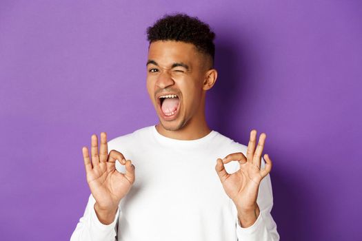 Cheeky african-american man winking, ensure you in something, showing okay signs in approval, standing pleased over purple background.