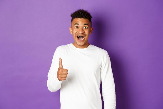 Portrait of amazed african-american man, showing thumbs-up and looking excited, recommend and praise good product, standing over purple background.