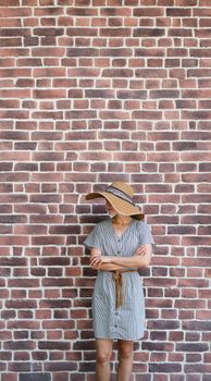 Summer style. Portrait of a beautiful young woman in summer dress and hat standing on red brick wall background