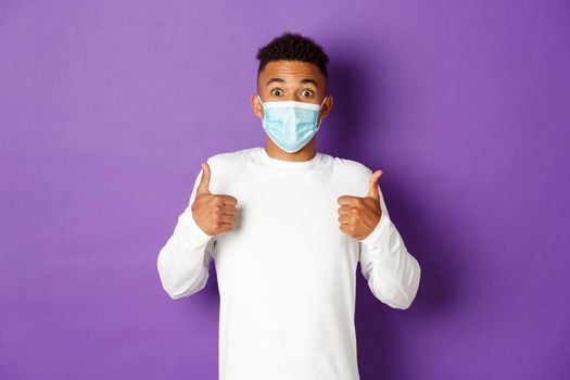 Concept of covid-19, pandemic and social distancing. Amazed african-american guy in medical mask, showing thumbs-up in approval, like something, standing over purple background.