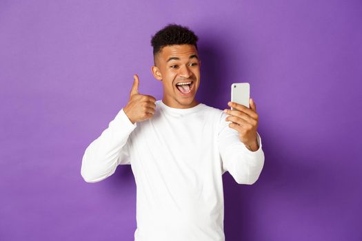 Image of smiling african-american male blogger, showing thumbs-up at mobile phone camera, recommend something, standing with smartphone over purple background.