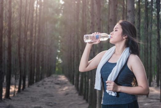 Beautiful young woman drinking water bottle after exercise fitness, in nature park forest