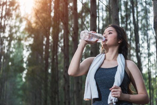 Beautiful young woman drinking water bottle after exercise fitness, in nature park forest