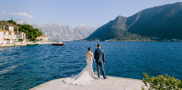 Bride and groom stand on the pier holding hands and look at the sea. Back view. High quality photo