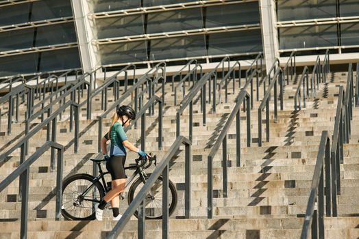 Young professional female cyclist in cycling garment and protective gear standing on the steps with her bike while exercising outdoors. Sports concept. Side view