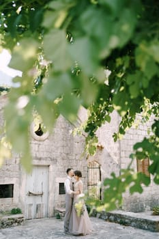 View through the foliage of a tree at the bride and groom hugging near the stone building. High quality photo