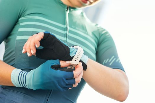 Cropped shot of professional female cyclist in sportswear putting on smart watch and cycling gloves while getting ready for training. Sports, extreme and active lifestyle concept