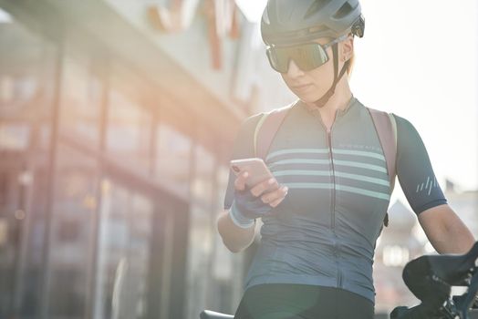 Female cyclist in protective gear using smartphone while riding bicycle in city center. Sportswoman training, exercising outdoors. Sports, active lifestyle concept