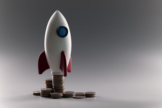 Toy rocket standing on piles of coins on gray background closeup. Startup in business concept
