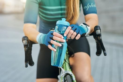 Cropped shot of professional female cyclist holding water bottle, resting, standing with her bike outdoors on a daytime. Active lifestyle, sports concept
