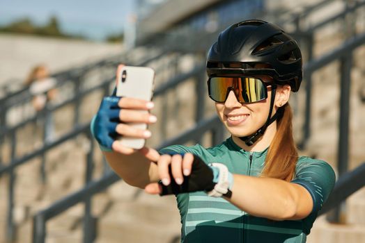 Portrait of professional female cyclist in cycling garment and protective gear smiling while taking selfie using smartphone, resting after riding bicycle in city center. Sports, technology concept