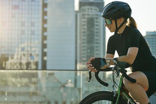 Smiling sportswoman in black cycling garment and protective gear having a rest, admiring cityscape while riding bike, training outdoors on a sunny day. Active lifestyle, sports concept