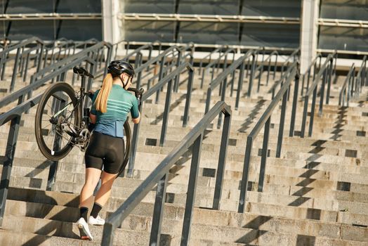 Full length shot of professional female cyclist in cycling garment and protective gear with bicycle on her shoulder walking up steps, training outdoors. Sports, active lifestyle concept. Rear view