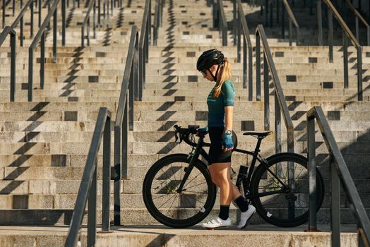 Full length shot of professional female cyclist in cycling garment and protective gear standing on the steps with her bike after having a training, riding in city center. Sports concept. Side view