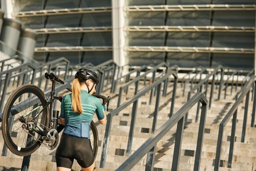 Rear view of female cyclist with bicycle on her shoulder walking up steps, having training in city. Sports, active lifestyle concept