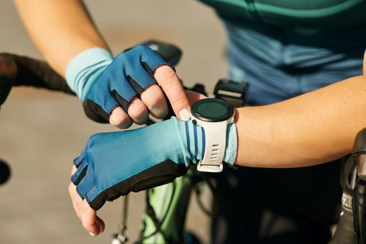 Close up shot of hands of professional female cyclist in cycling garment using smartwatch, checking results after having a training, riding bicycle on a sunny day. Sports, extreme, active lifestyle