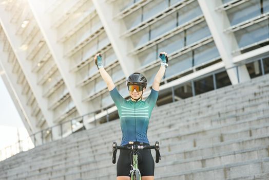 Excited professional female cyclist in cycling garment raised her arms, smiling at camera while standing with her bike outdoors on a daytime. Active lifestyle, sports concept