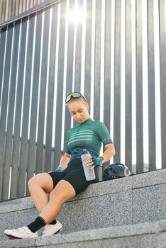 Young woman, professional cyclist in sporty shorts, with bottle of drink resting on the steps in the city background. Urban lifestyle, riding bicycle, sports concept