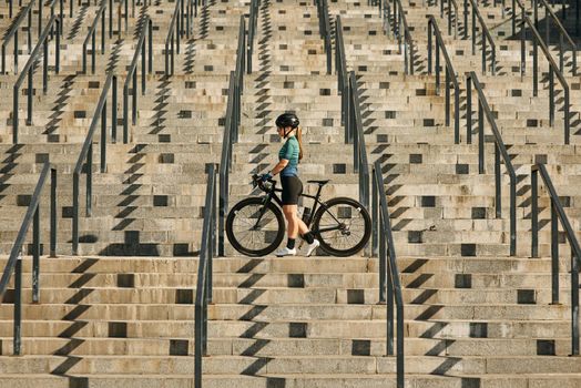 Young professional female cyclist in cycling garment and protective gear standing on the steps with her bike after having a training, riding in city. Sports concept. Side view