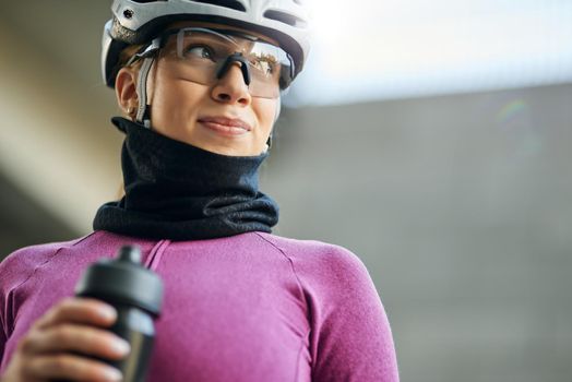 Portrait of cute professional female cyclist wearing pink suit and neck warmer looking away, holding water bottle while getting ready for training, standing outdoors on a daytime. Sports concept