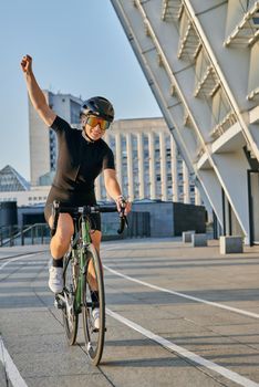 Excited professional female cyclist in black cycling garment and protective gear smiling, raising her arm, looking satisfied with the result of her training. Sports, active lifestyle concept