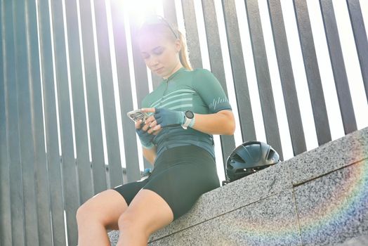 Low angle view of young woman, professional cyclist in sportwear using smartphone while resting on the steps in the city. Urban lifestyle, riding bicycle, sports concept